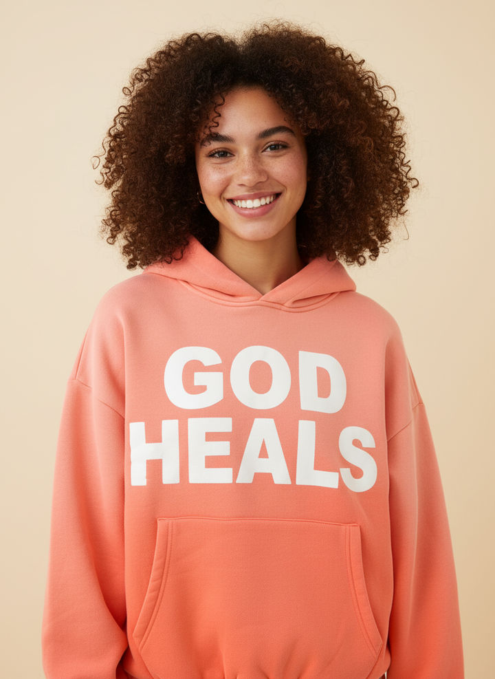 Hoodie ‘God Heals’ voor dames, christelijke streetwear-mode tegen een lichtoranje achtergrond.
